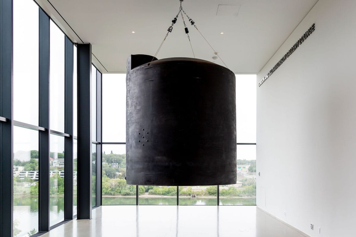 Postcommodity, Let Us Pray For the Water Between Us, 2020, 2,200-gallon polyethylene hazmat chemical storage container, brushless linear motor, leather mallet, wood, steel, aircraft cable, algorithmic composition. Originally commissioned by Minneapolis Institute of Art. Installation view, Time Holds All the Answers, Remai Modern, 2021. Photo: Carey Shaw.