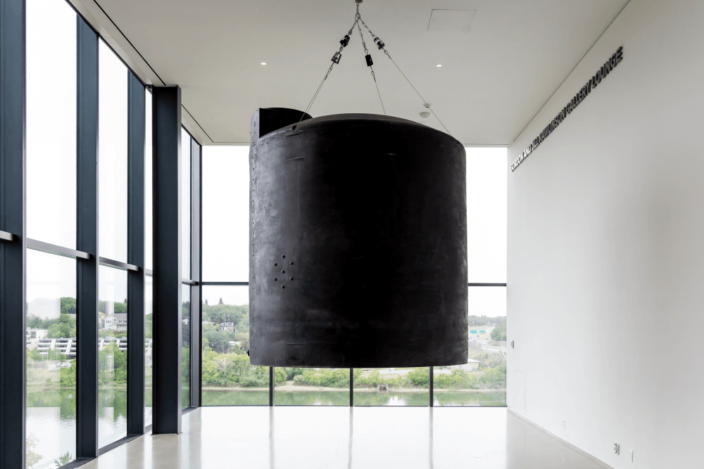 Postcommodity, Let Us Pray For the Water Between Us, 2020, 2,200-gallon polyethylene hazmat chemical storage container, brushless linear motor, leather mallet, wood, steel, aircraft cable, algorithmic composition. Originally commissioned by Minneapolis Institute of Art. Installation view, Time Holds All the Answers, Remai Modern, 2021. Photo: Carey Shaw.