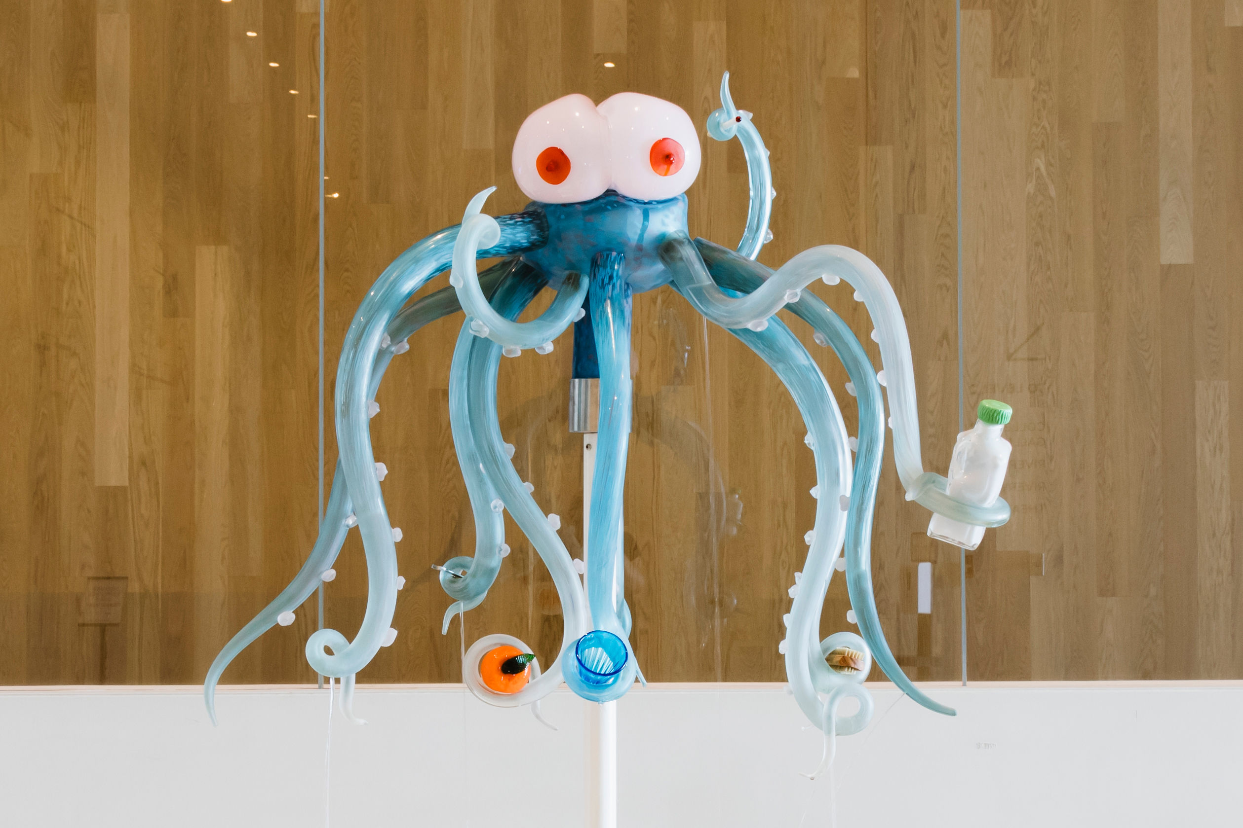 A Venetian glass sculpture depicts an octopus-like creature with breasts for a head. The creature is grasping various items in its tentacles including an orange and a glass.