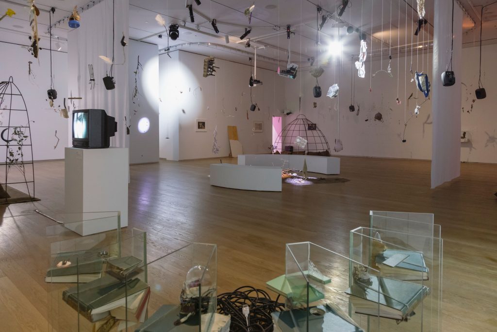A gallery space shows various works by Laure Prouvost, including installations, video works on hung sheets, and numerous mobiles.