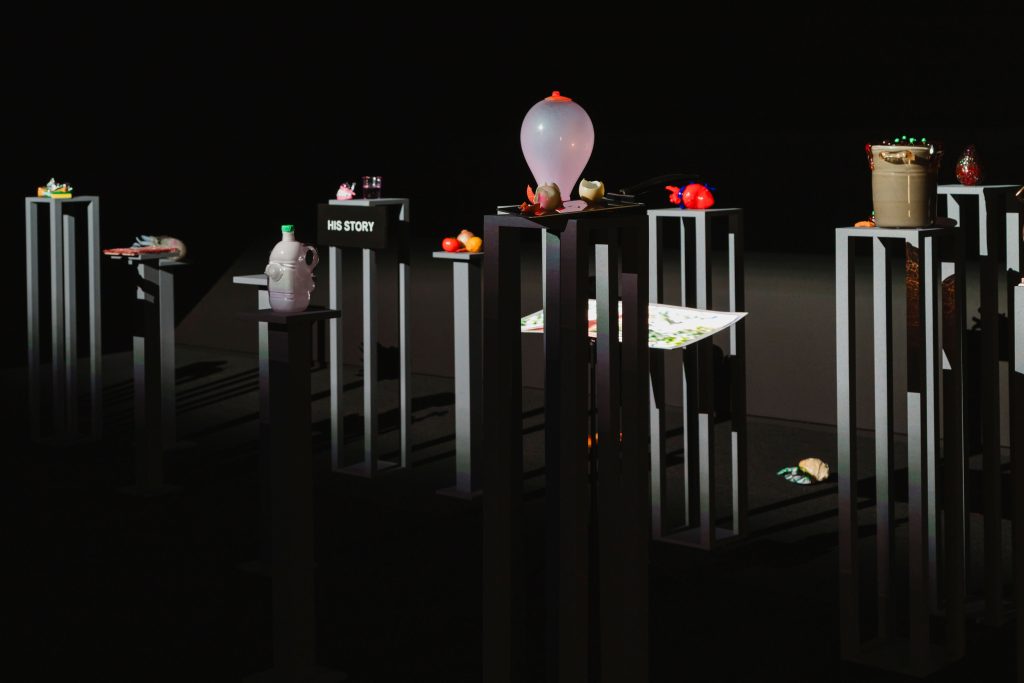 A grouping of tall, narrow black platforms hold various objects, including fruits and vegetables, a model heart and dentures. They are displayed in a black room and are illuminated by lights off camera.