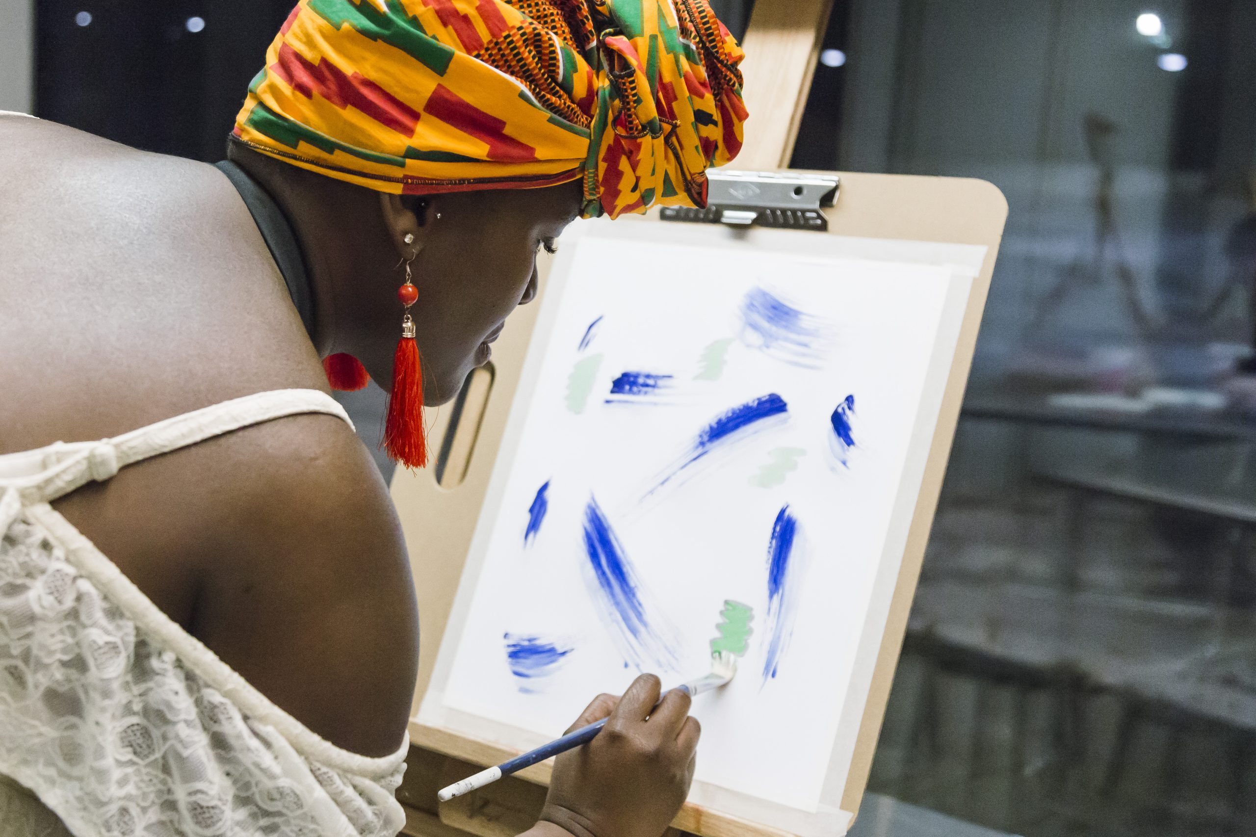 A woman wearing a brightly coloured head wrap adds blue and green marks to a canvas.