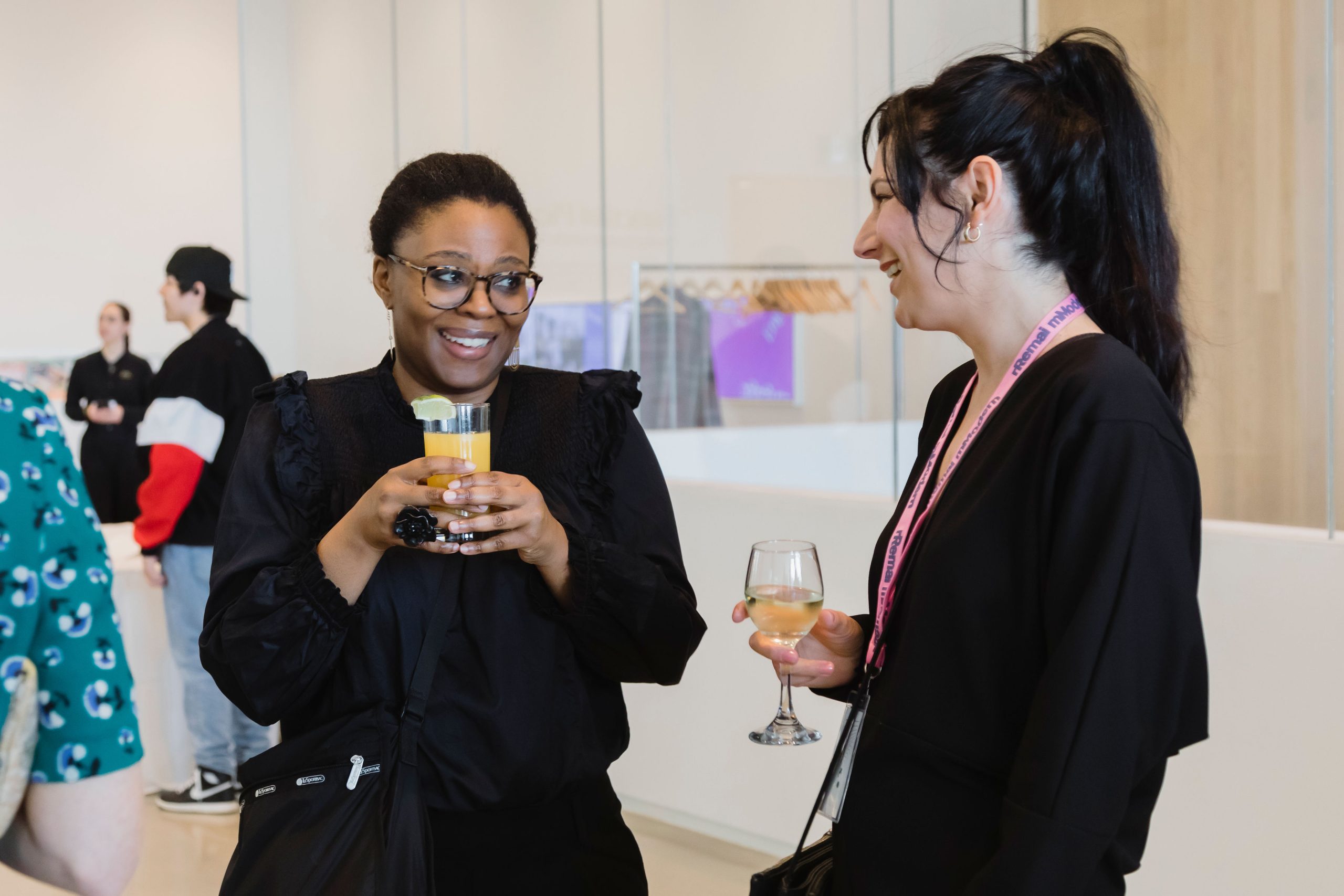 Two people talk with each other, smiles on their faces and drinks in hand. Others are socializing in the background.