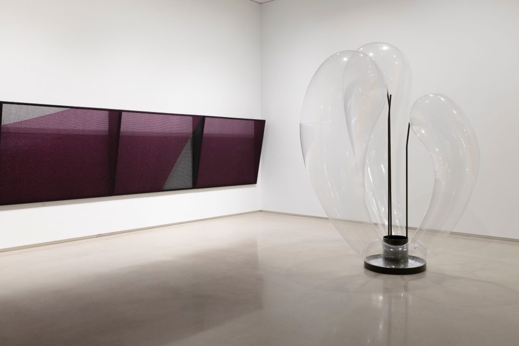 An inflatable sculpture with three bulbous arms stands on the floor next to a woven work hung on the wall.