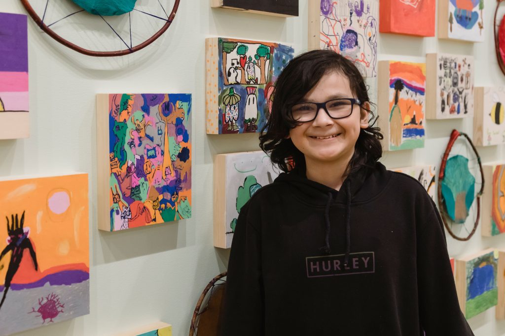 A child smiles next to their artwork in the exhibition All Aboard: Everyone an Artist.