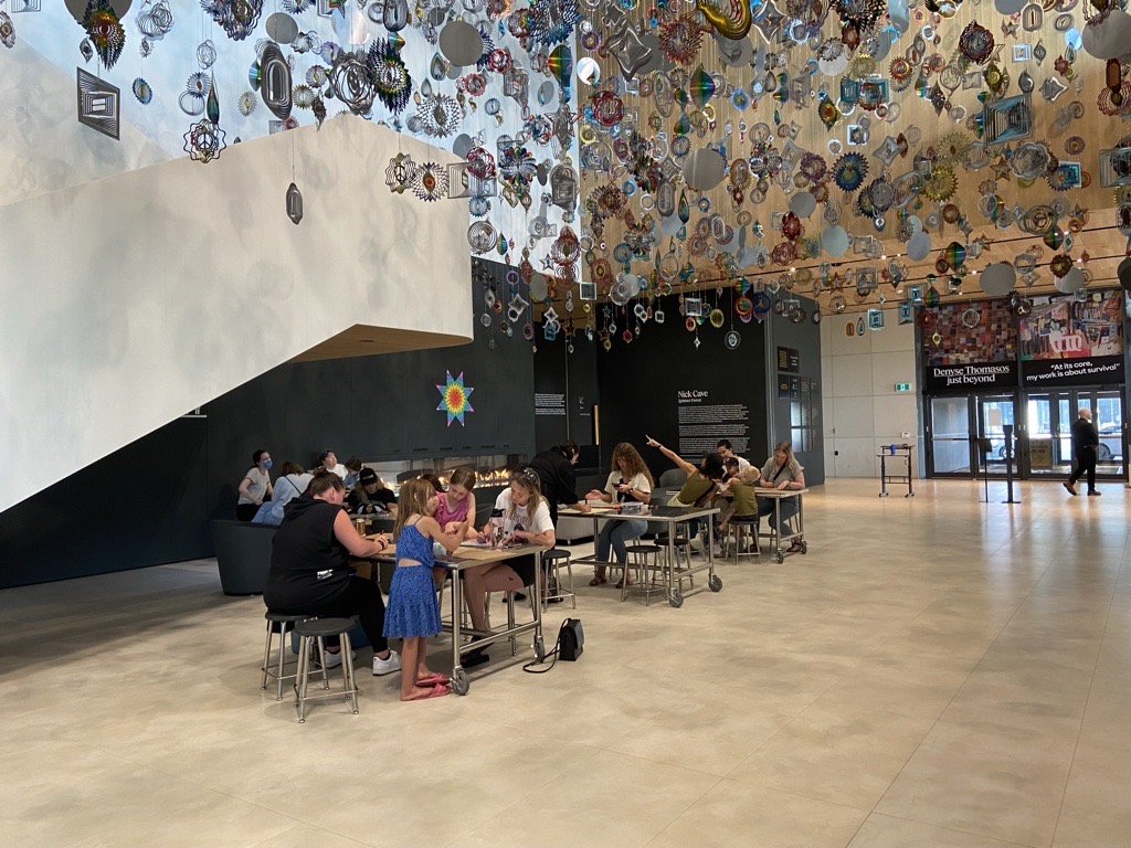 People sit at tables together, making Pride Parade signs in Remai Modern's atrium.
