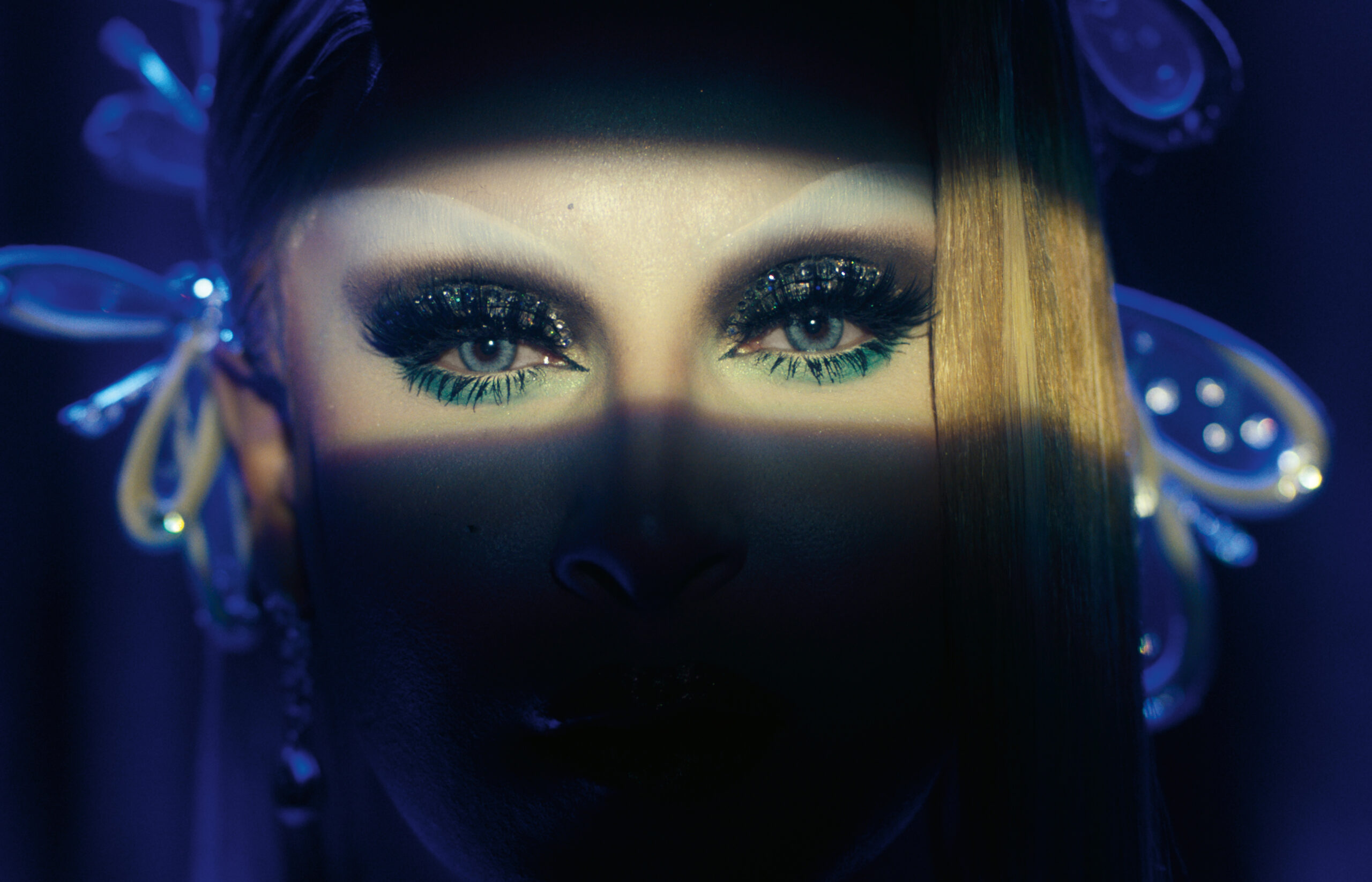 A person looks at the camera, their face covered in shadow except their eyes, which are covered in glamorous makeup.