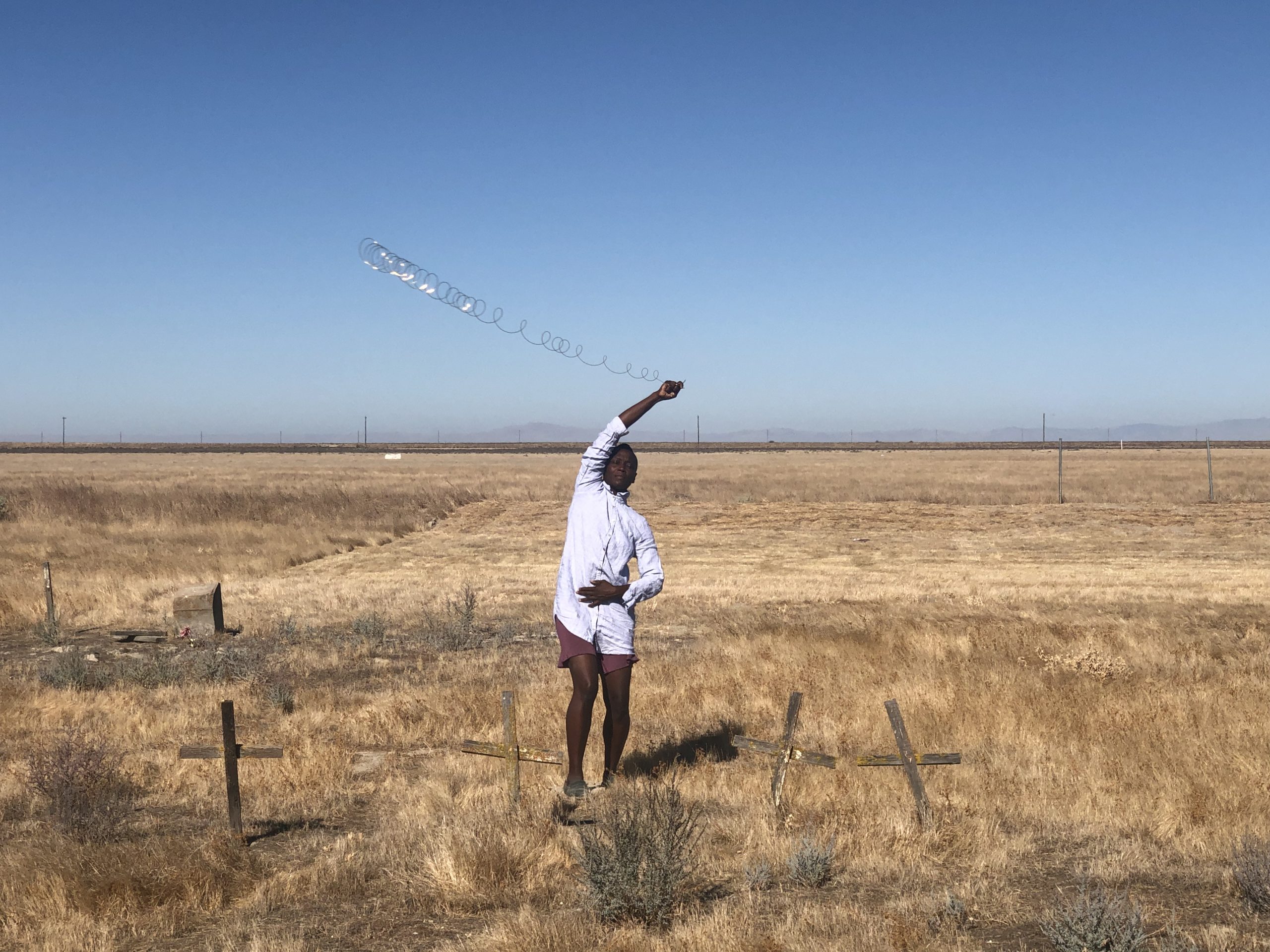 A photo of artist taisha paggett standing in a field covered in dry grass and a few weathered wooden crosses. The artist is wearing a light blue, long-sleeved button-up shirt and brown shorts and is swinging a coiled piece of wire around her head.