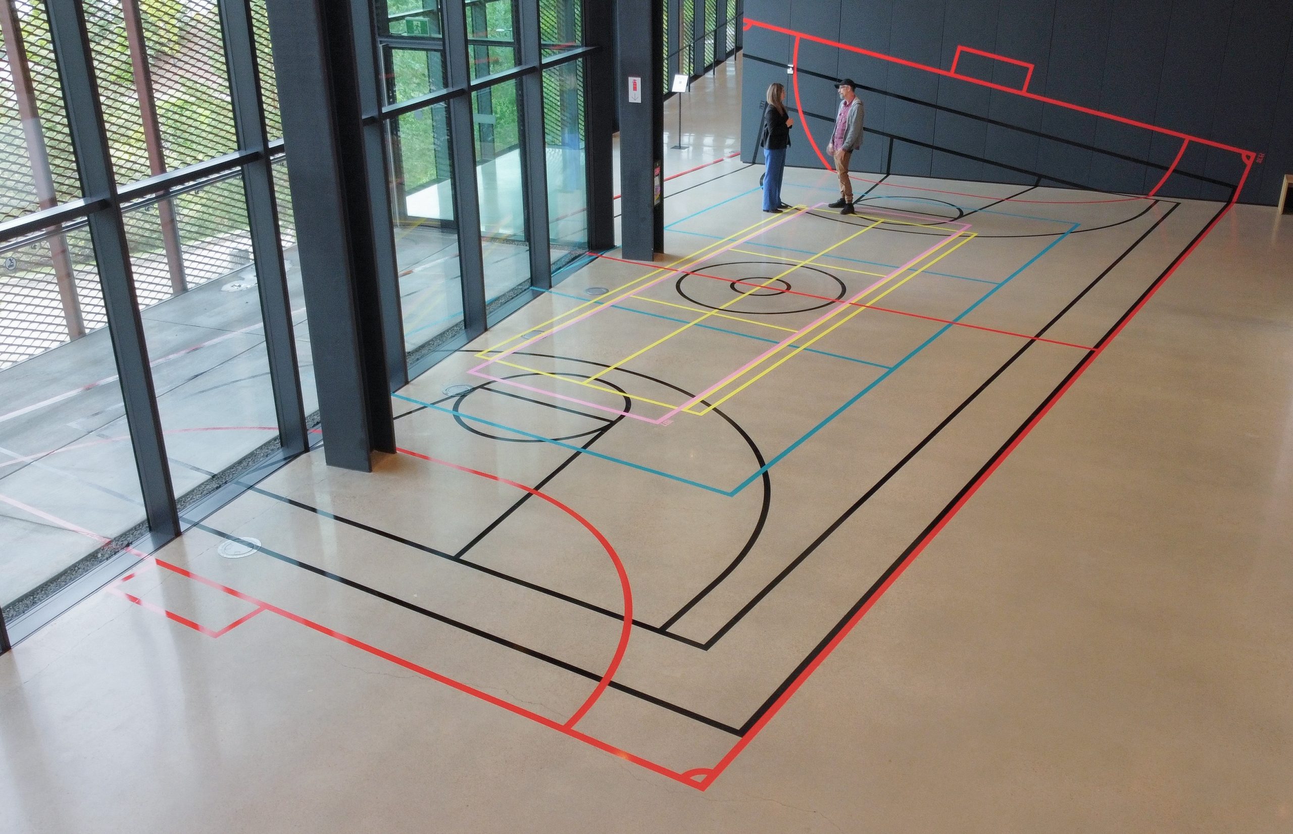 An indoor installation featuring a sports court layout made of colorful tape lines, designed to resemble a hybrid of different sports fields. Two people are seen standing and conversing on the court, which is situated in a modern, glass-walled space, allowing natural light to fill the room.