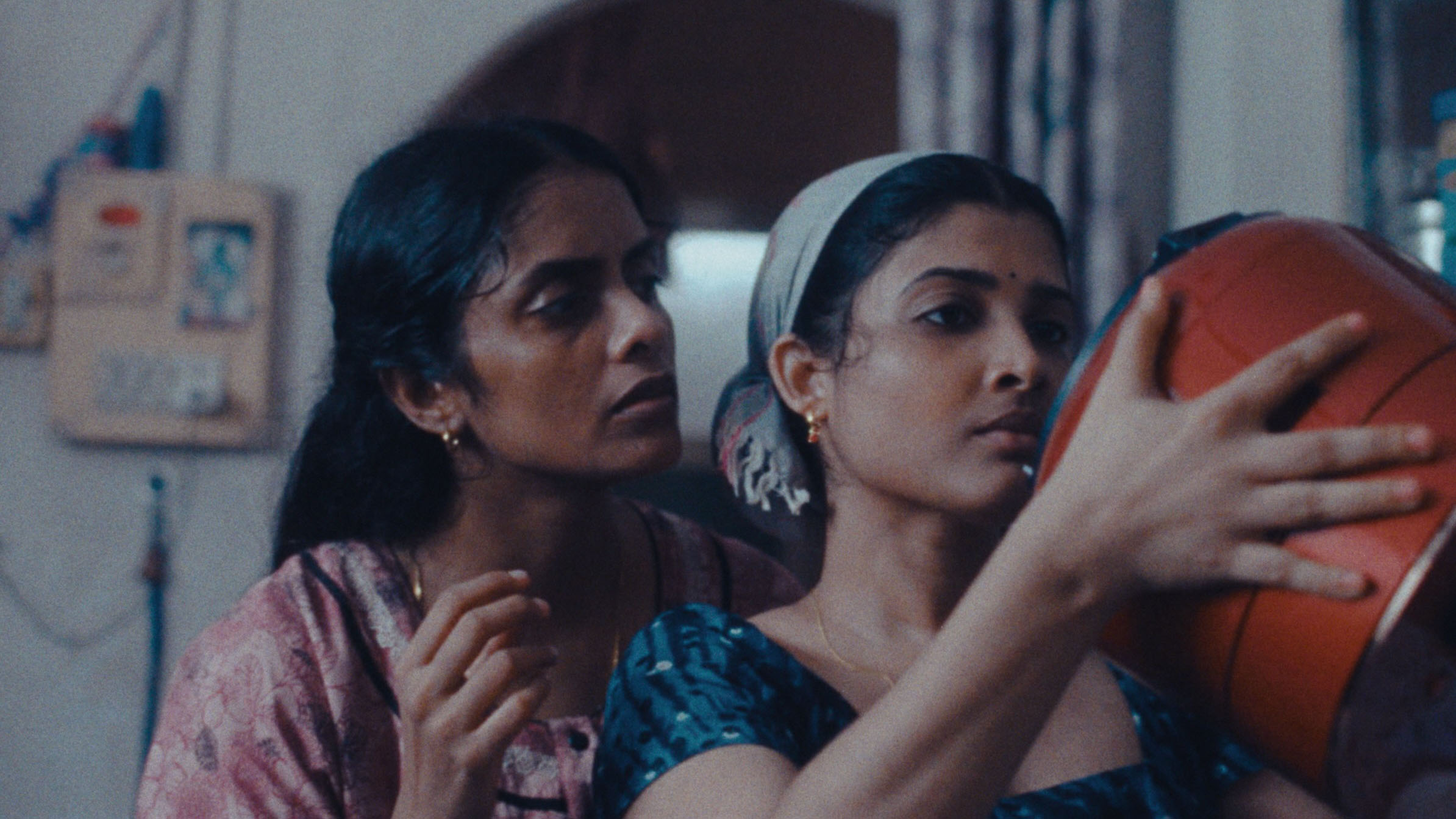 Two women stand closely together in a dimly lit room, focused on examining a red cooking pot. The woman on the right, wearing a teal blouse and a headscarf, holds the pot with both hands, concentrating intently. The woman on the left, dressed in a pink patterned top, looks over her shoulder with a serious expression. The setting suggests a domestic space, and the scene conveys a sense of quiet tension or urgency.