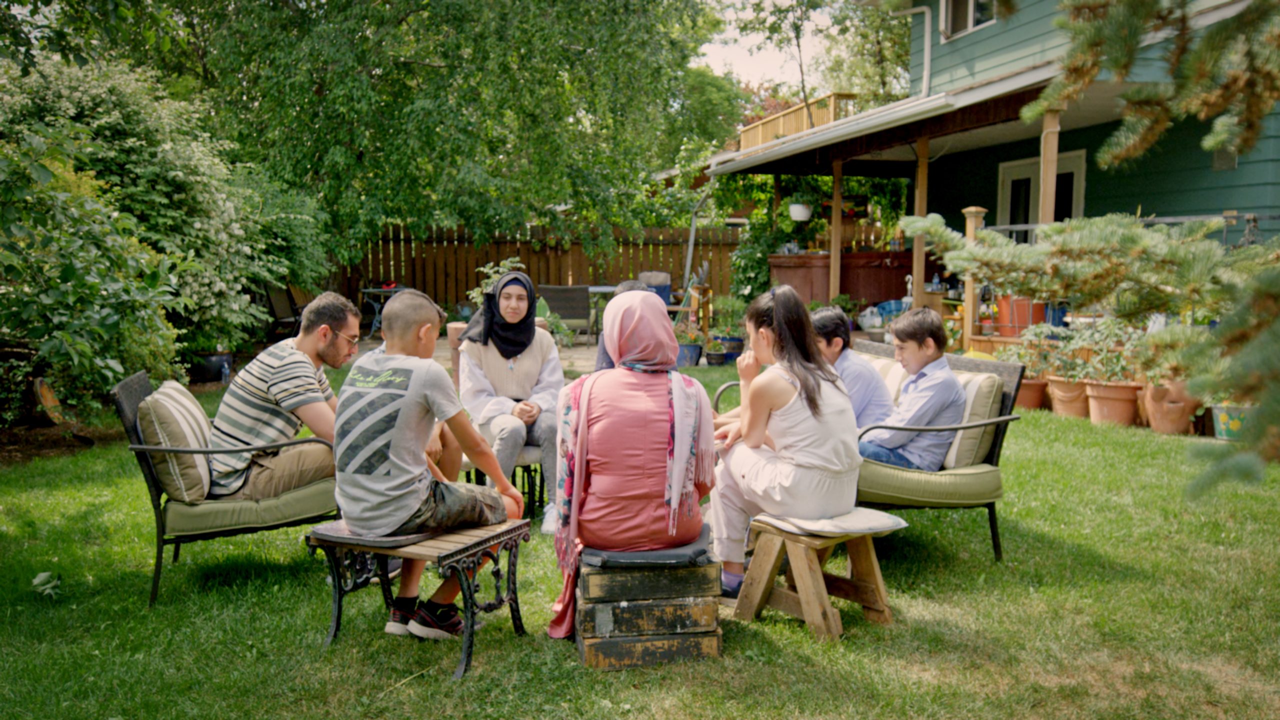 A group of people sit in a circle on chairs and stools in a lush backyard, engaged in what appears to be a serious or heartfelt conversation. The group includes men, women, and children, with some individuals wearing headscarves. The setting is peaceful, with green grass, potted plants, and a wooden fence and house in the background. Sunlight filters through the trees, giving the moment a quiet, reflective tone.