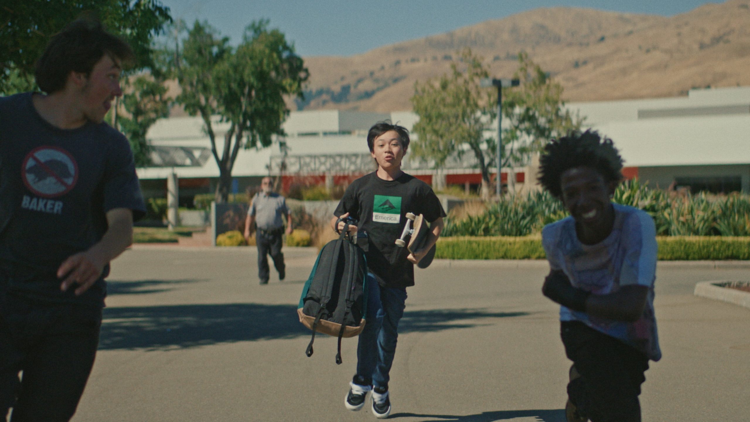Three teenage boys run toward the camera in a school parking lot on a sunny day. The boy in the center carries a backpack and a skateboard, wearing a black T-shirt. Another boy on the left looks back as he runs, and the boy on the right smiles broadly. In the background, a security guard appears to be chasing them, with low, rolling hills and a school building behind.