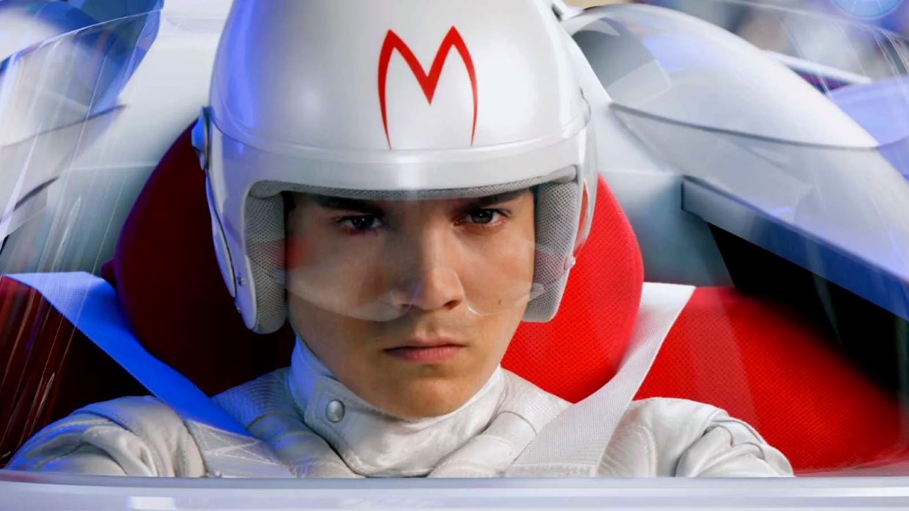 A young man in a white racing suit and helmet sits in a futuristic race car, staring ahead with intense focus. His helmet has a bold red "M" on the front, and he is seated in a red bucket seat. The image has a glossy, high-contrast look, suggesting a stylized or CGI-enhanced film scene.