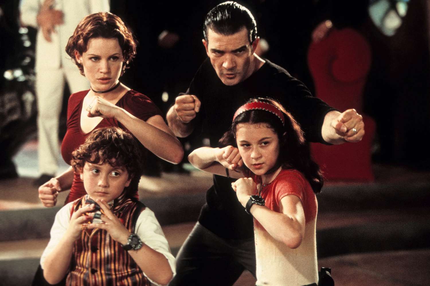 A family of four stands in martial arts stances, looking determined and ready for action. The father, dressed in black, stands behind his wife and two children, all with fists raised. The mother wears a red top and choker, the daughter has a red headband and matching shirt, and the younger son wears a striped vest over a white shirt. Each of them wears a high-tech gadget on their wrist. The scene has a stylized, adventurous feel, suggesting a spy or action-themed story.