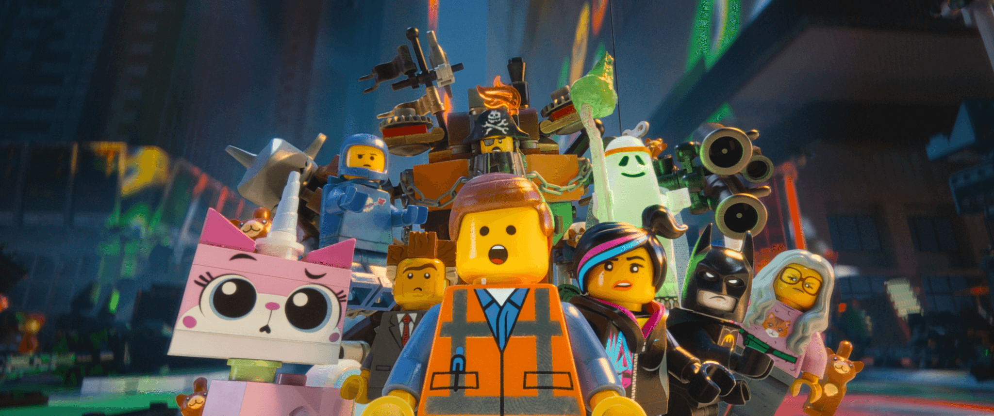A group of LEGO minifigure characters stands in the foreground of a colorful, neon-lit LEGO city, all looking forward with surprised or determined expressions. Emmet, wearing an orange safety vest, is front and center with his mouth open. Behind him are Wyldstyle with streaked hair, a pink cat-unicorn named Unikitty, a classic space astronaut in a blue suit, a pirate with mechanical parts, a friendly ghost, LEGO Batman in his black cowl, and an older woman figure in glasses. They appear ready for action, surrounded by vivid urban lights and towering buildings.
