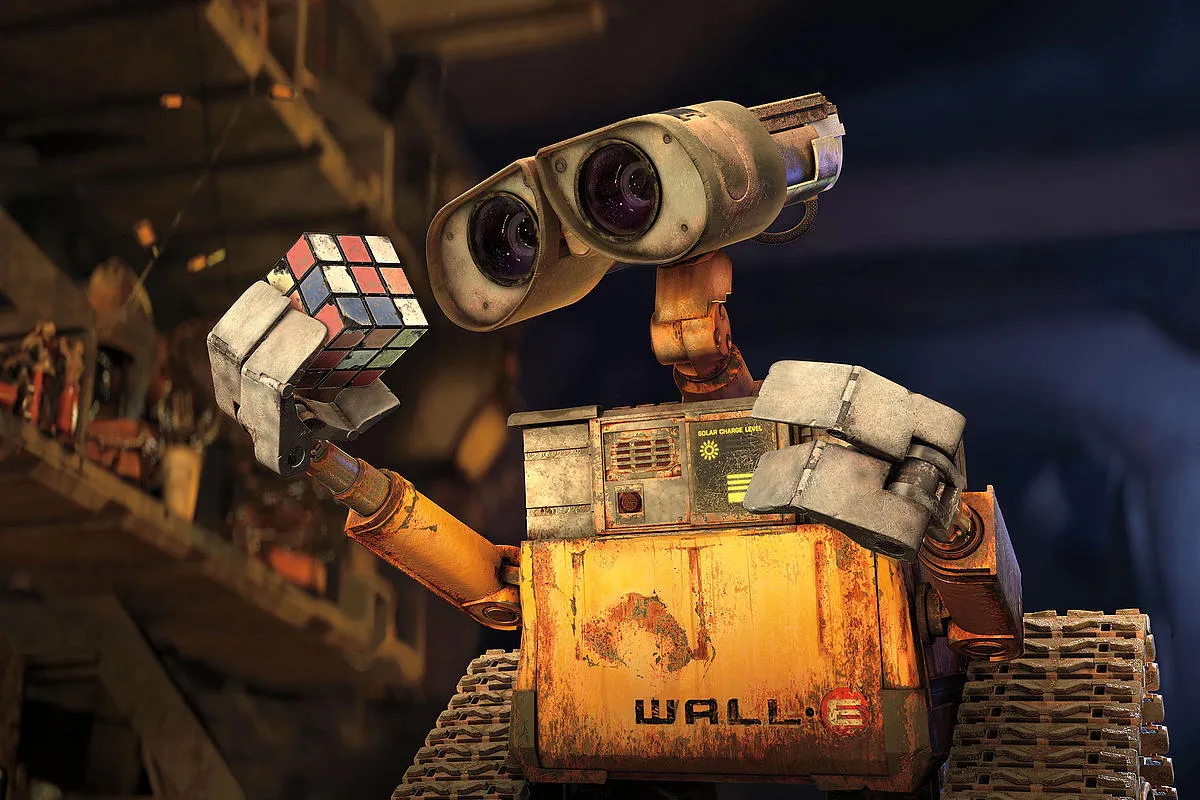A rusty, expressive robot named WALL·E holds up a partially solved Rubik's Cube with curiosity. He has large, binocular-like eyes and treads for movement. The background is dimly lit, showing shelves cluttered with various objects, suggesting a post-apocalyptic setting. The robot's chest panel displays a solar charge level.