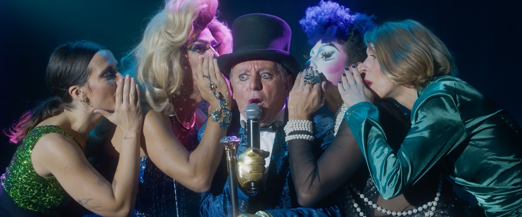 A flamboyantly dressed performer in a blue sequined suit, top hat, and gold glove stands center stage holding a microphone, eyes wide in surprise. Four elaborately styled individuals—wearing glittery makeup, wigs, and colorful costumes—stand close on either side, dramatically whispering into the performer's ears. The lighting is theatrical, casting a blue and pink glow that heightens the sense of drama and campy spectacle.