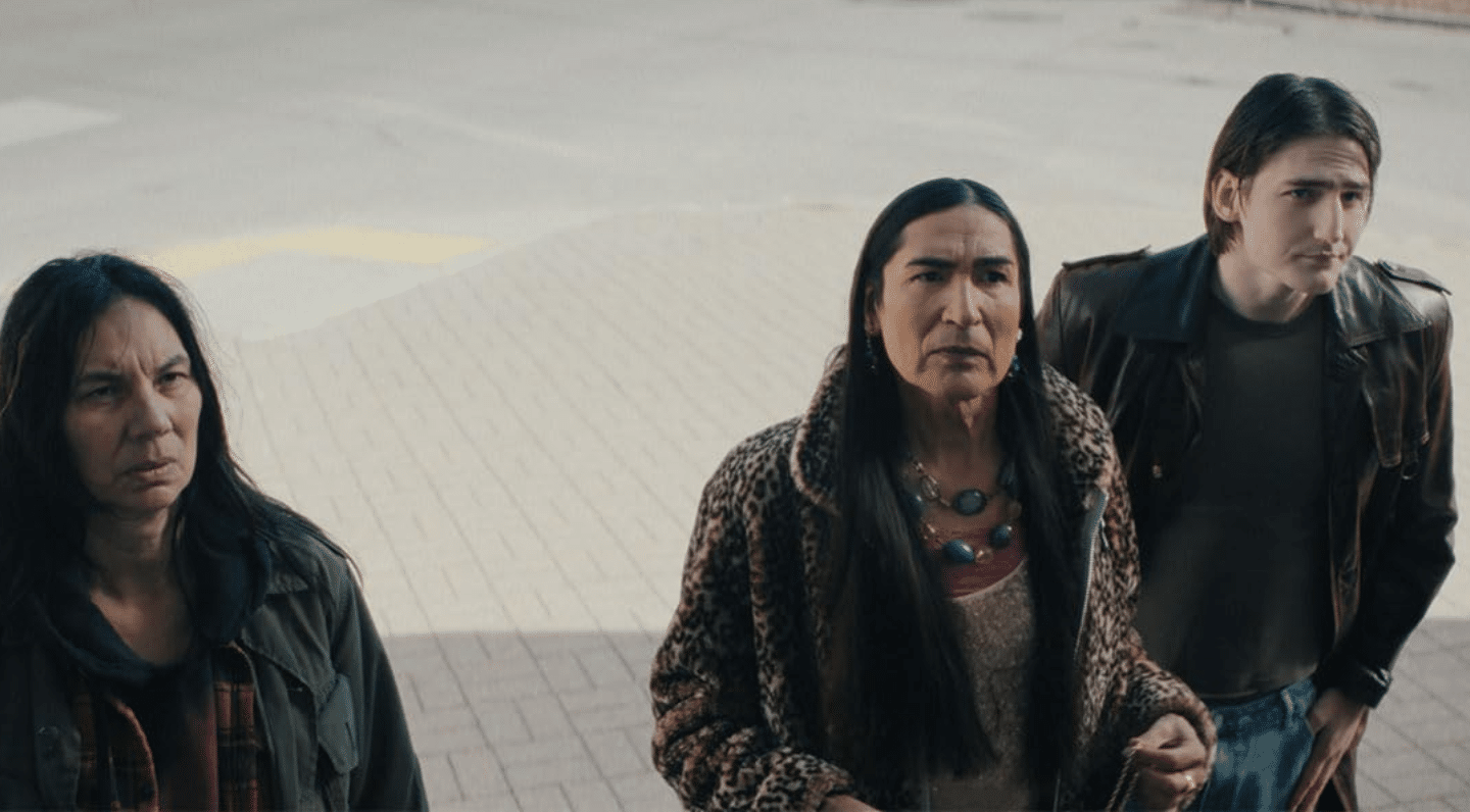 Three people stand side by side outside on a paved surface, looking upward with serious or curious expressions. The person in the center wears a leopard print coat and a beaded necklace, with long straight hair, while the individuals on either side wear darker, casual clothing—one in a flannel-lined jacket, the other in a leather jacket. The lighting is soft and natural, suggesting a contemplative or pivotal moment.