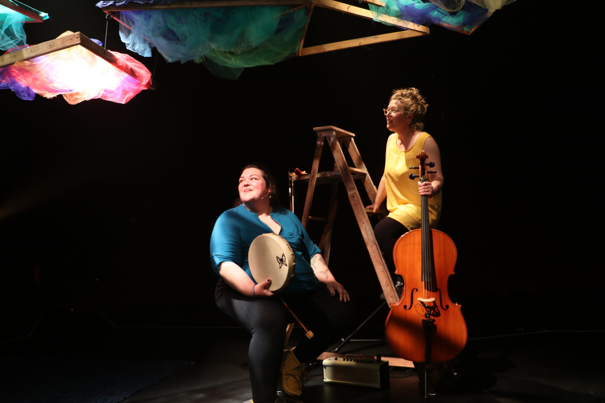 Two musicians perform on a dimly lit stage with colorful fabric decorations overhead. One musician, wearing a blue top, sits on a stool holding a hand drum and smiling. The other musician, wearing a yellow top, leans against a wooden ladder while holding a cello. A small amplifier is placed on the floor nearby, contributing to the performance setting.