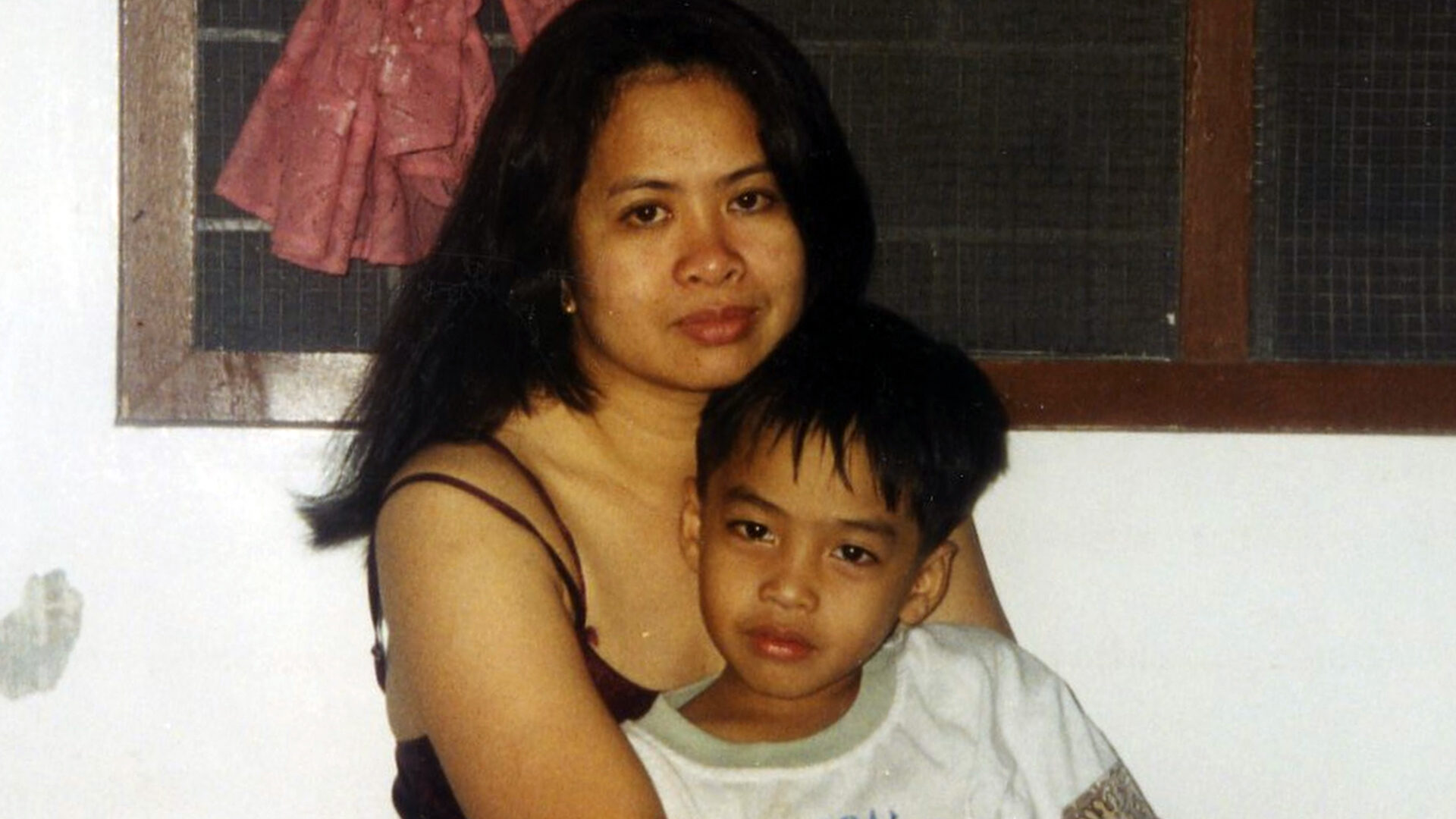 A woman and a young boy sit closely together indoors, both looking directly at the camera with calm, neutral expressions. The woman has long dark hair and wears a thin-strapped top, while the boy rests against her in a white shirt. Behind them is a simple wall with a wooden-framed window and a pink garment hanging. The image has a personal, intimate, and slightly nostalgic feel, resembling a candid family photo.