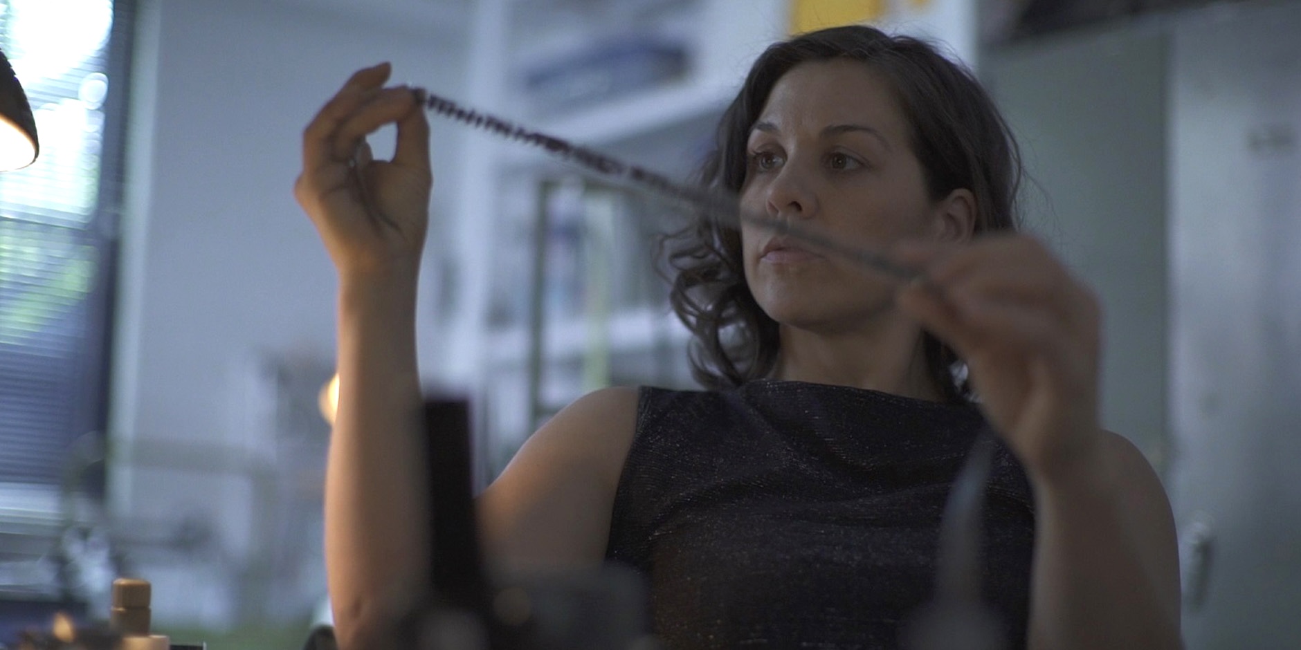 A woman with wavy dark hair sits in a dimly lit room, carefully examining a strip of film held between both hands. Her expression is focused and contemplative. The background is softly blurred, with shelves and various objects suggesting a creative or archival workspace. Light from a nearby lamp casts a soft glow, emphasizing the detail of the filmstrip.