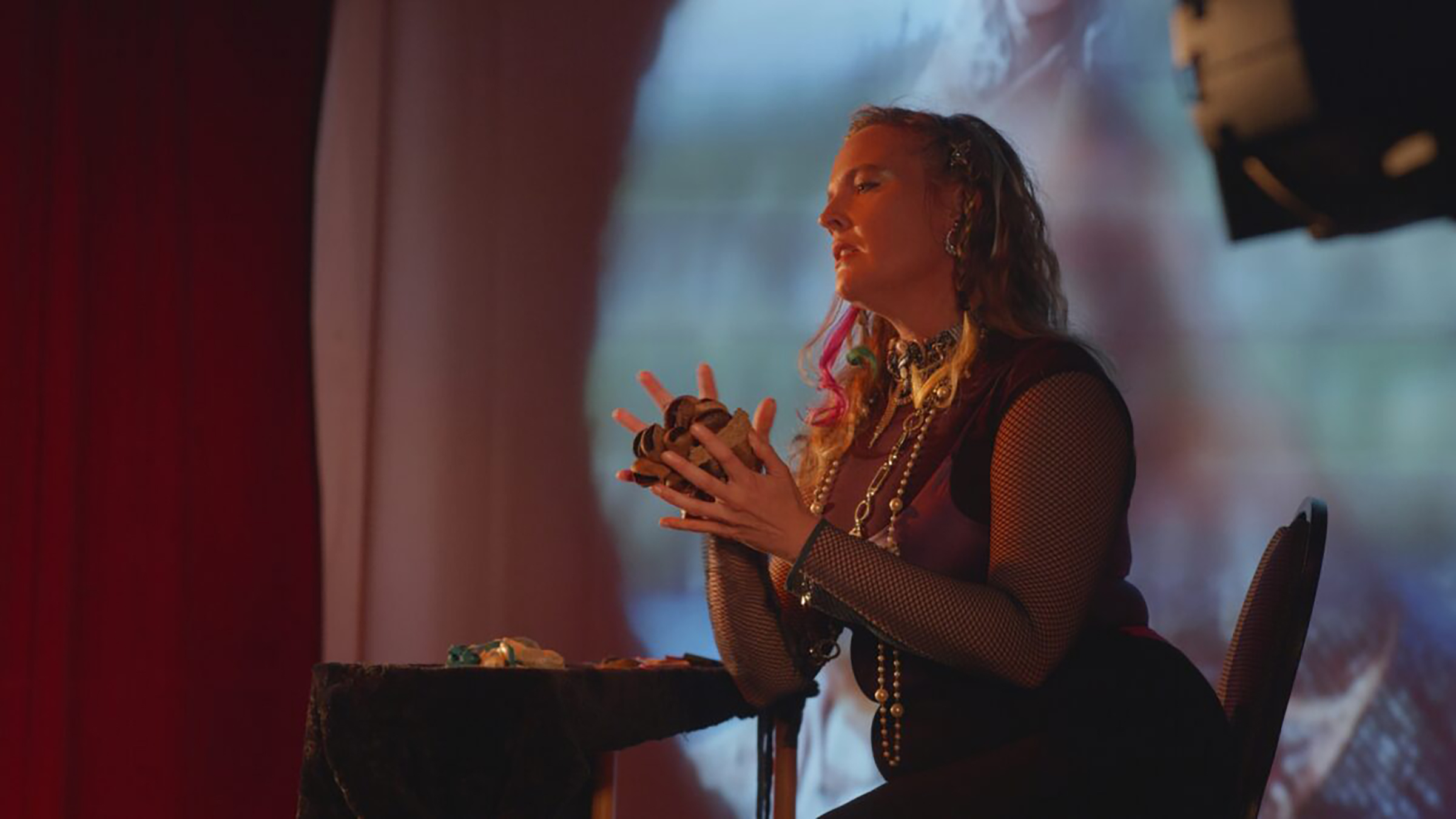 A person with long wavy hair and colorful highlights sits on a stage, illuminated by warm lighting. They wear a dark sleeveless top over a mesh shirt, layered necklaces, and hold an object in their hands, possibly a natural item like driftwood or shells. A table covered in velvet and adorned with small items sits in front of them. Behind them, a projected image adds a dreamy, surreal atmosphere to the scene.