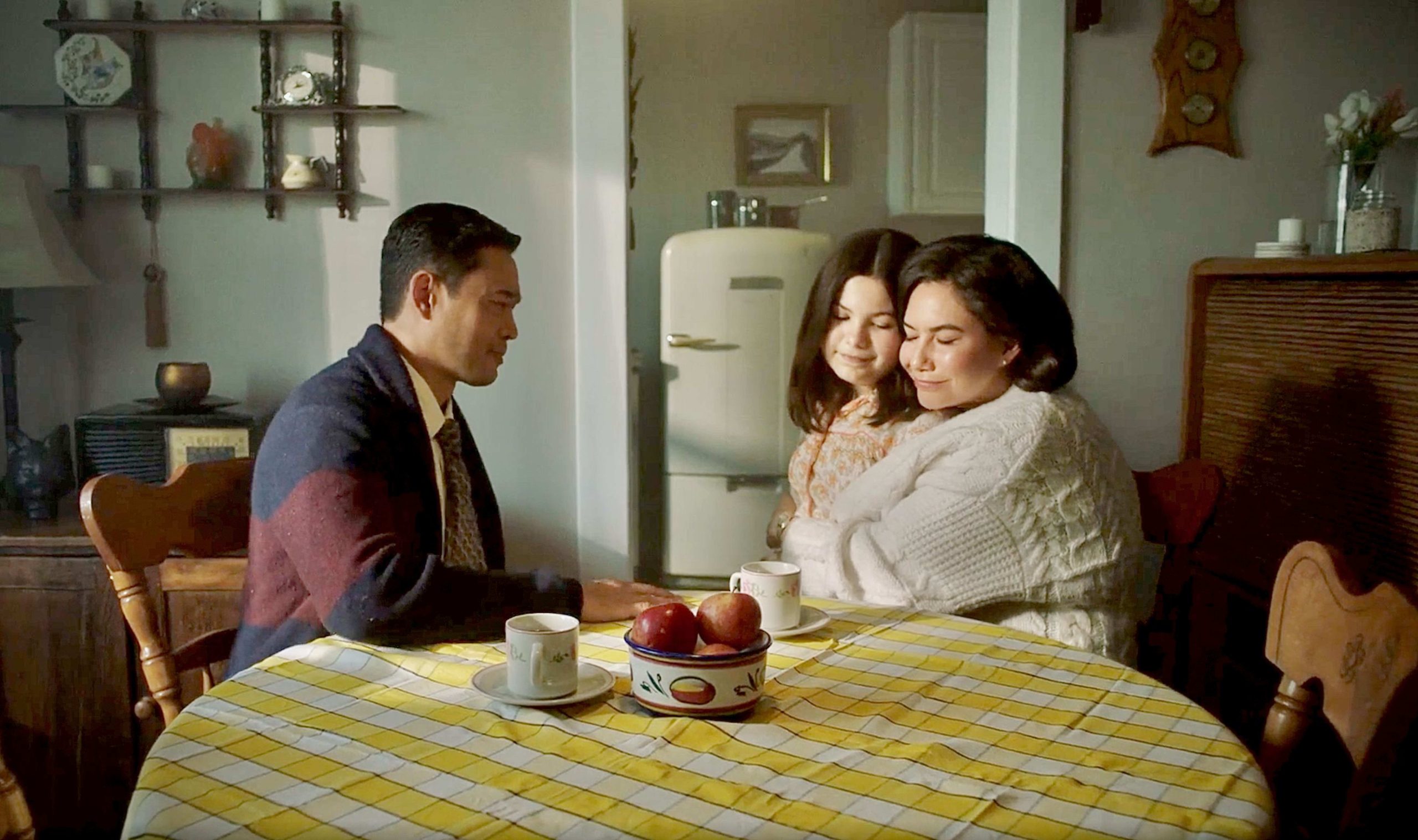 A warm, intimate scene of a family seated around a kitchen table with a yellow checkered tablecloth. A man in a suit and cardigan sits on the left, facing a woman and a young girl on the right, both wearing cozy sweaters and leaning into each other with peaceful expressions. A bowl of apples and cups of coffee or tea sit in the foreground. The background features vintage kitchen decor, including an old-style refrigerator and wooden shelves, evoking a nostalgic, mid-20th-century atmosphere.