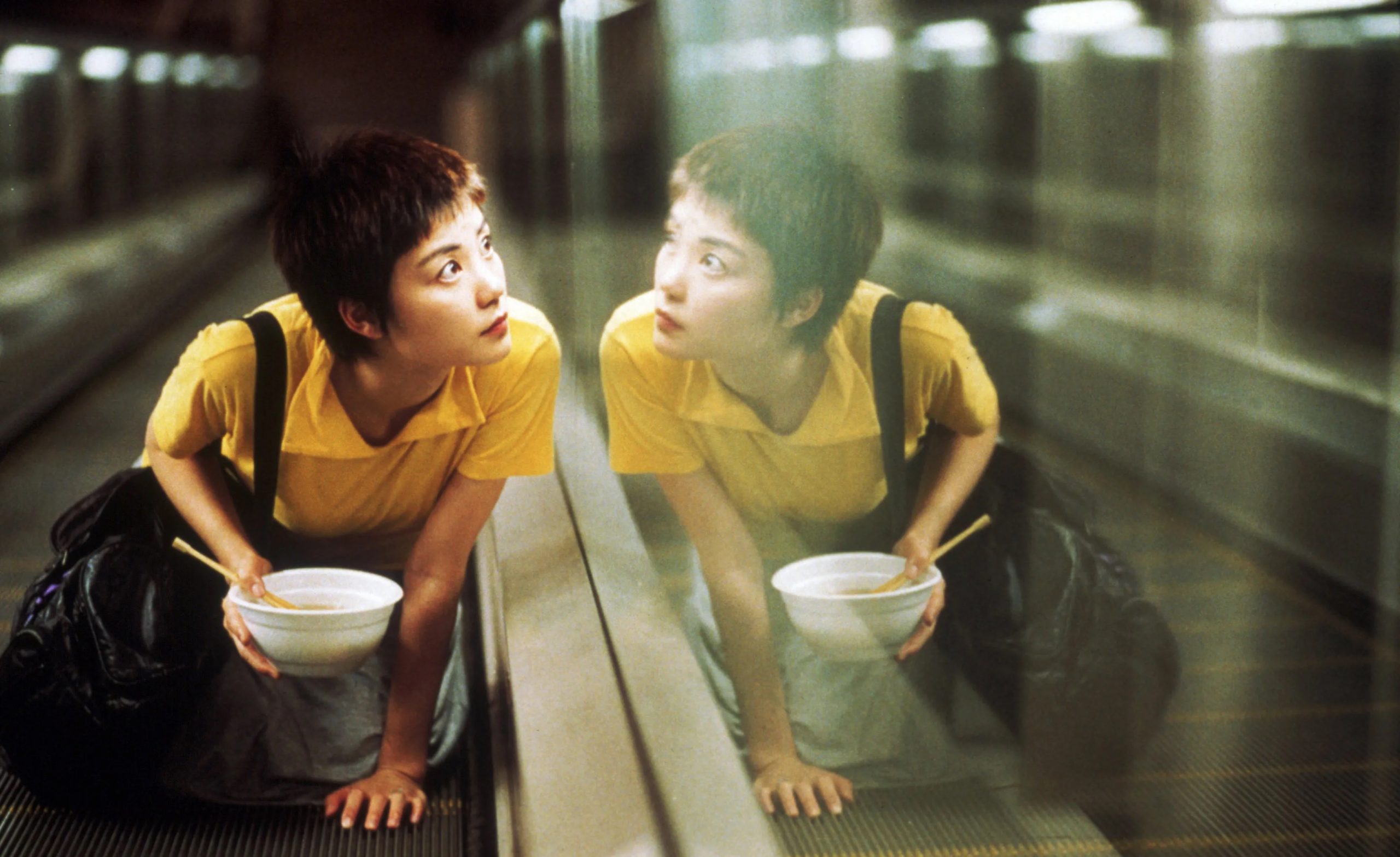 A young woman with short hair and a yellow shirt kneels on a moving walkway, holding a white bowl and looking intently at her reflection in the glass wall beside her. She has a bag slung over one shoulder, and the scene is bathed in soft, diffused lighting, creating a dreamy, introspective mood. The setting appears to be an empty, dimly lit subway or airport corridor.