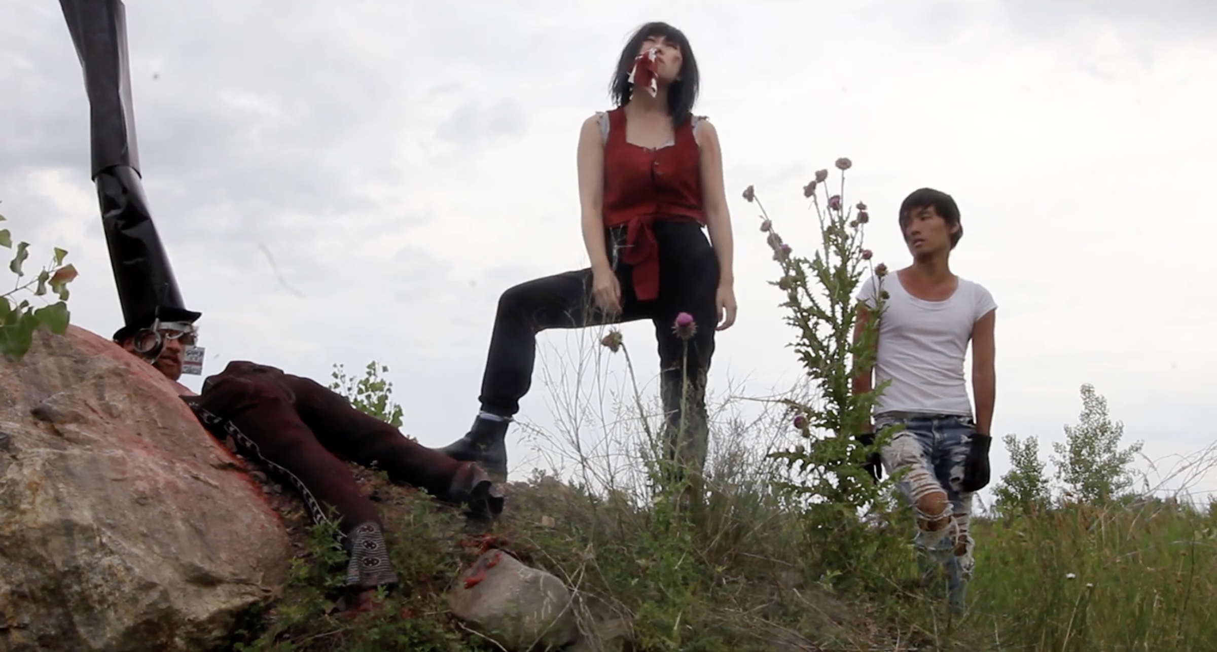 In a grassy, overcast landscape, a woman in a red top and black pants stands with one foot on a rock, blood on her mouth and a ribbon hanging from it. Next to her, a young man in a torn white shirt and ripped jeans looks on passively, wearing a single black glove. Behind the woman lies a man slumped against the rock, dressed in a steampunk-style outfit with goggles and a tall black top hat. The scene is eerie and surreal, evoking a post-apocalyptic or avant-garde atmosphere.