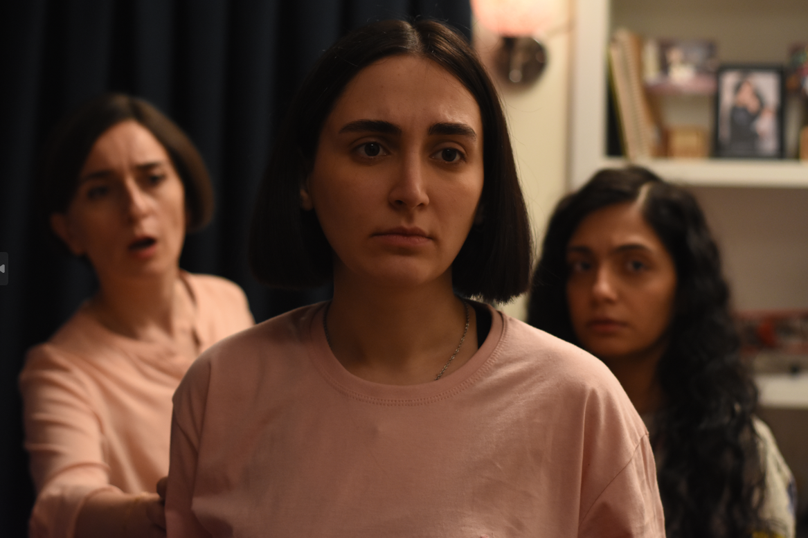 Three women stand closely together indoors, with serious and concerned expressions. The woman in the foreground wears a light pink shirt and stares ahead intently. Behind her, one woman appears to be speaking or reacting urgently, while the third watches quietly. The room is softly lit, with a bookshelf and framed photos in the background, creating a tense and emotionally charged atmosphere.