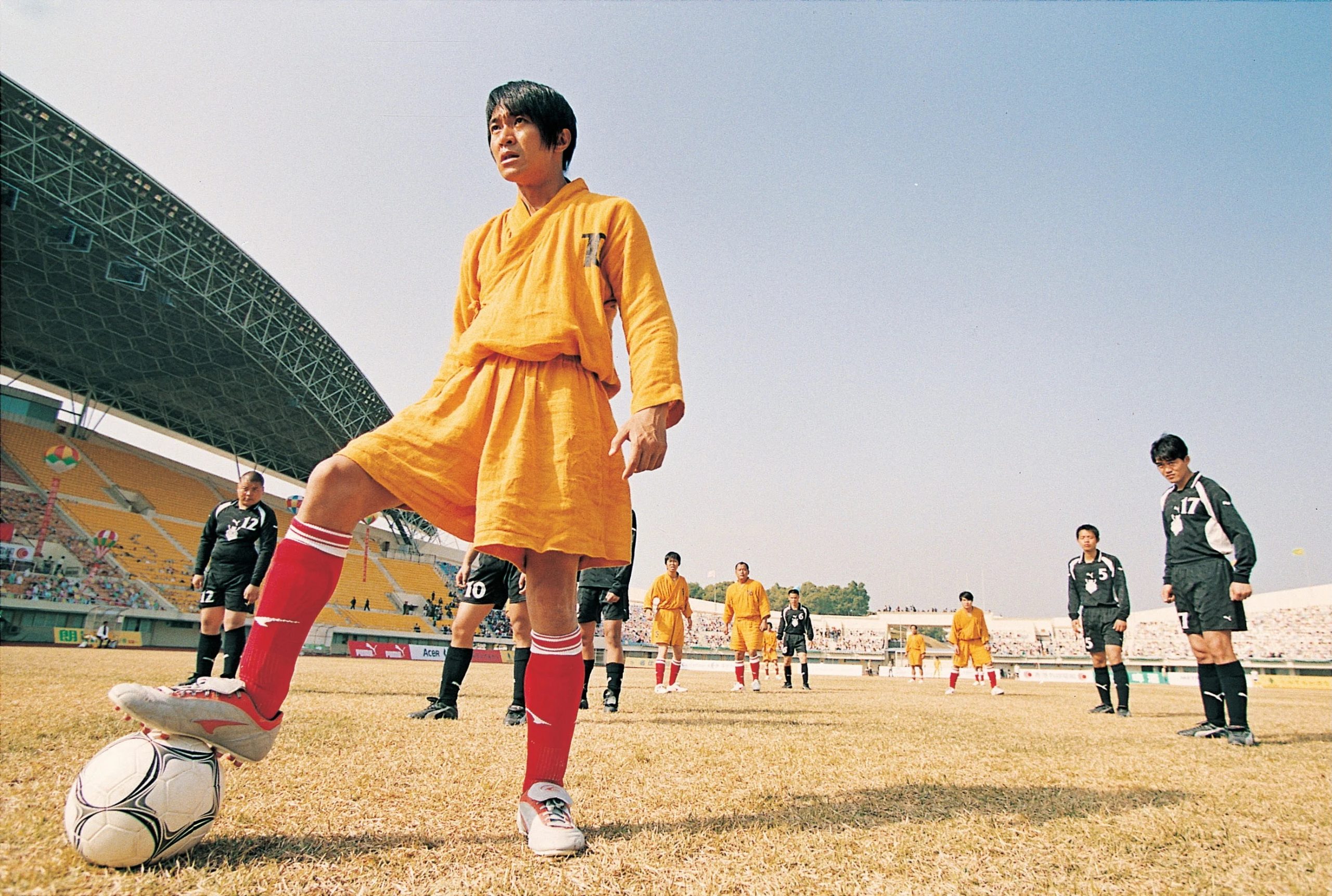 A soccer player in an orange monk-style robe and red knee-high socks stands confidently on a field, one foot resting on a soccer ball. Teammates in matching orange outfits and opponents in black uniforms are positioned behind him. The stadium is large and sunlit, filled with spectators. The scene combines elements of martial arts and sports, creating a dramatic and unconventional athletic moment.