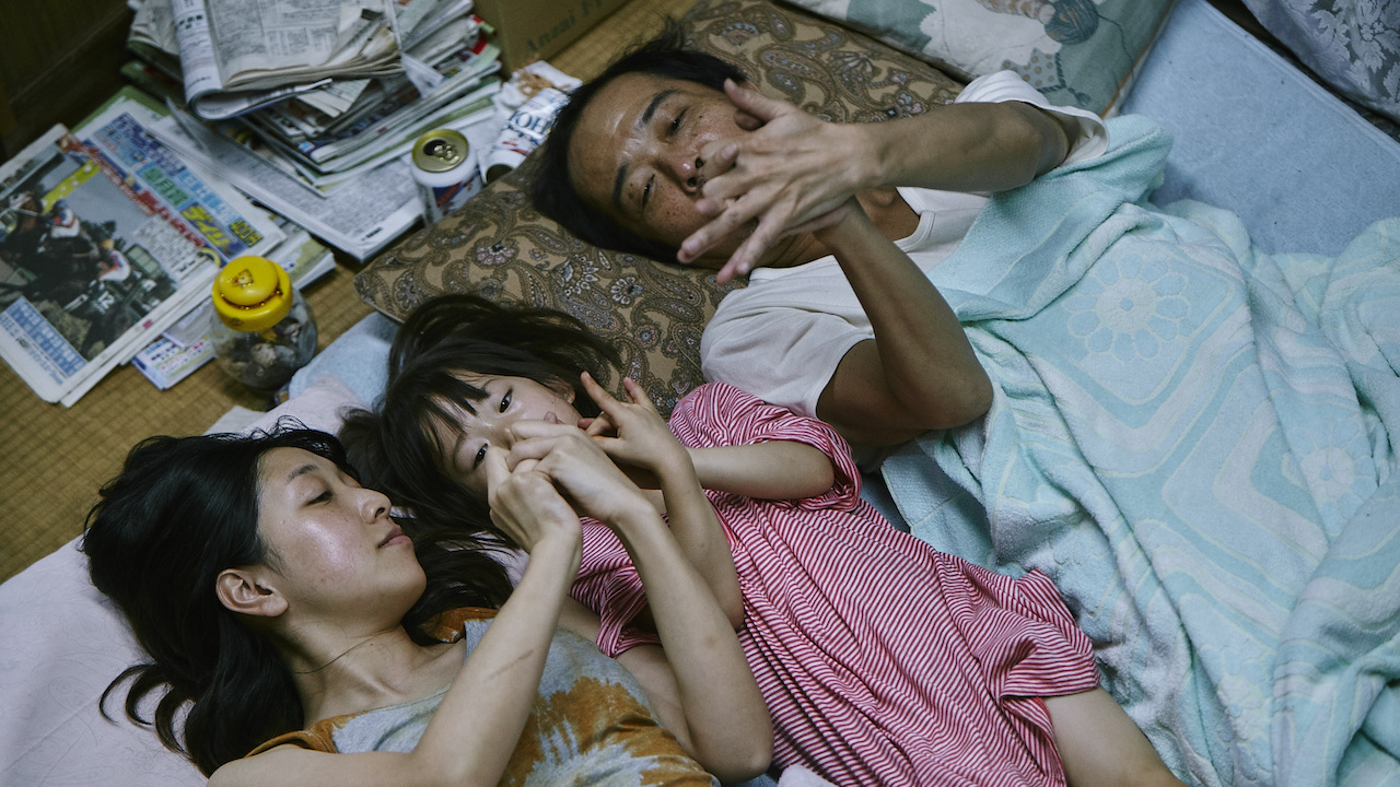 A man, woman, and young girl lie close together on thin bedding in a small, cluttered room, playfully linking fingers and gazing upward. The girl, dressed in a red striped outfit, lies between the two adults. Stacks of newspapers, a jar of coins, and miscellaneous items sit on the floor nearby, suggesting a humble living situation. Despite the modest surroundings, the moment feels warm and intimate, conveying a sense of familial closeness and quiet tenderness.