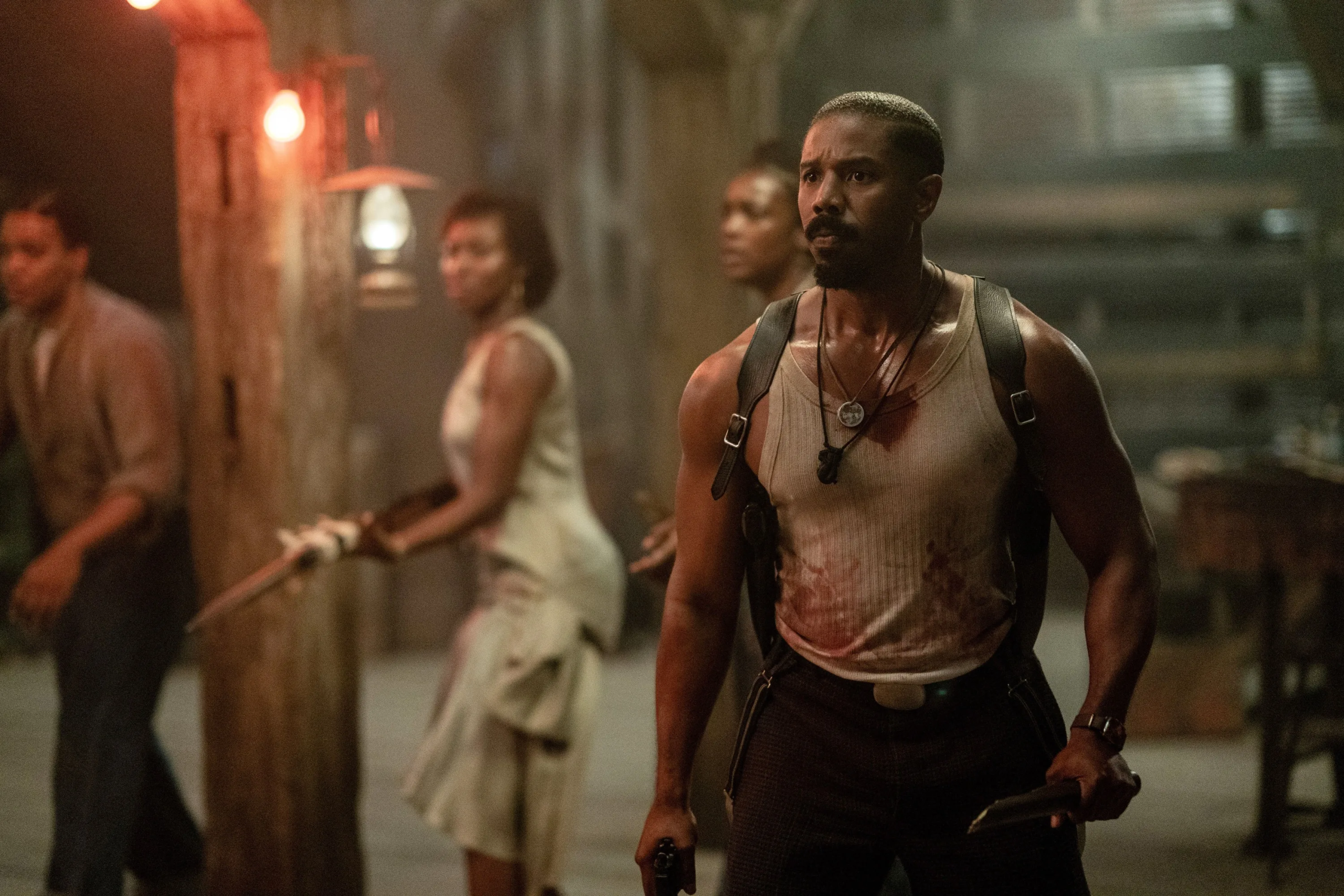 In a dimly lit, smoky interior, a muscular man stands in the foreground gripping a weapon, his sleeveless shirt stained and his expression tense and alert. He wears a harness and a pendant necklace, and his body language suggests he is bracing for danger. Behind him, several other people—also dirty and bloodied—hold makeshift weapons and look in different directions, as if surrounded or preparing to fight.