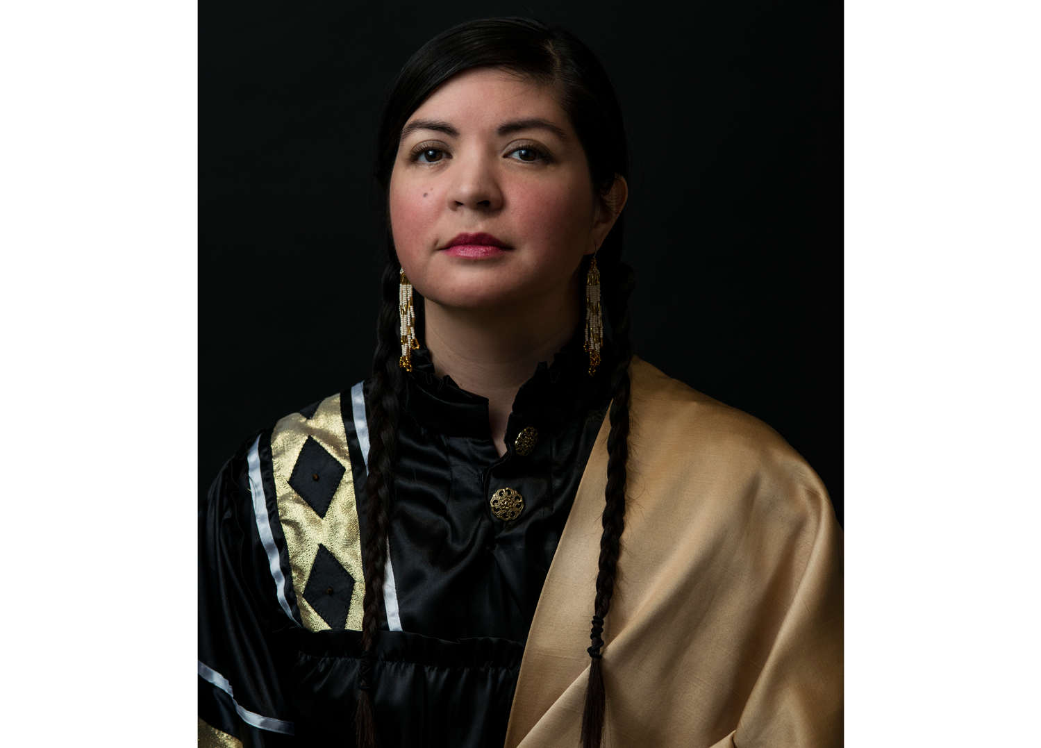 Studio portrait of a woman with two long braids, wearing a black satin dress with gold accents and a gold shawl, along with beaded earrings, photographed against a dark backdrop.